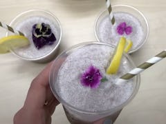 A woman's hand holds up a glass of violet lemonade with a flower in it, while two other glasses sit ...
