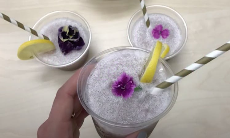 A woman's hand holds up a glass of violet lemonade with a flower in it, while two other glasses sit ...