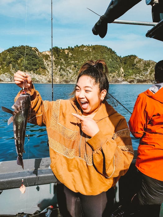 A woman in a yellow sweatshirt and leggings smiles and points to the fish she caught on a boat.