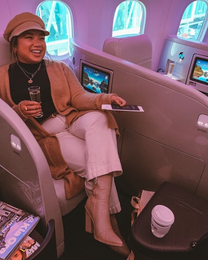 A woman wearing a long cardigan, white jeans, and booties smiles and sits in business class on an ai...