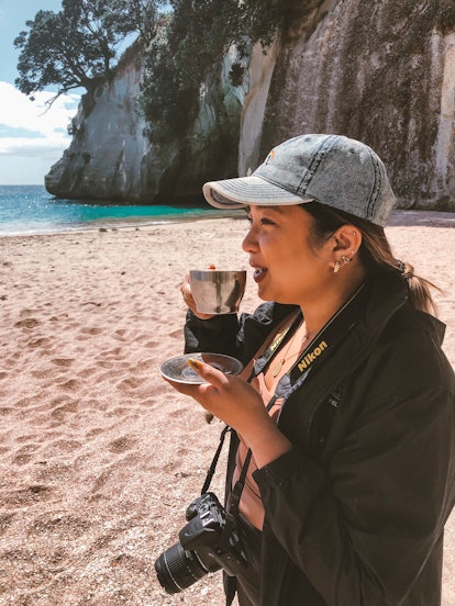 A woman in a baseball cap holds a cup of tea on a beach.
