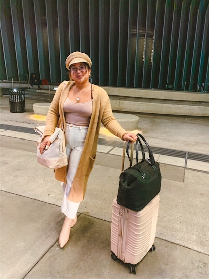 A woman wearing white pants, booties, a long cardigan and a hat poses with her rolling suitcase and ...