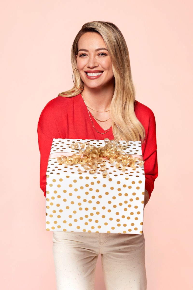 Hilary Duff in a coral sweater holding a white with gold polka dot present