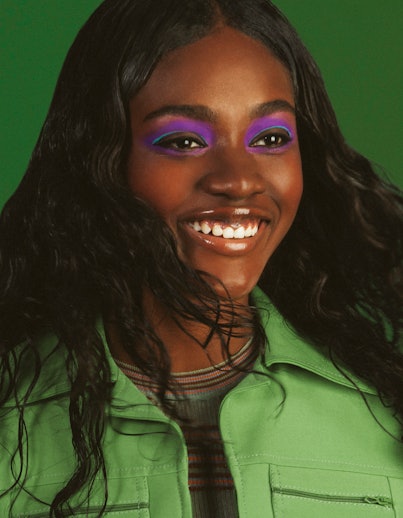 Zuri Tibby is smiling while wearing a green jumpsuit and purple eye shadow with a green background