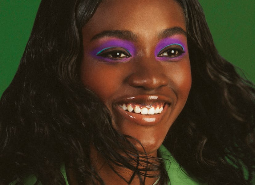 Zuri Tibby is smiling while wearing a green jumpsuit and purple eye shadow with a green background