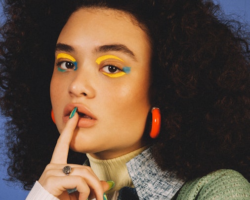 Diana Veras with yellow and blue eye make-up showing off her long nails and orange hoop earrings