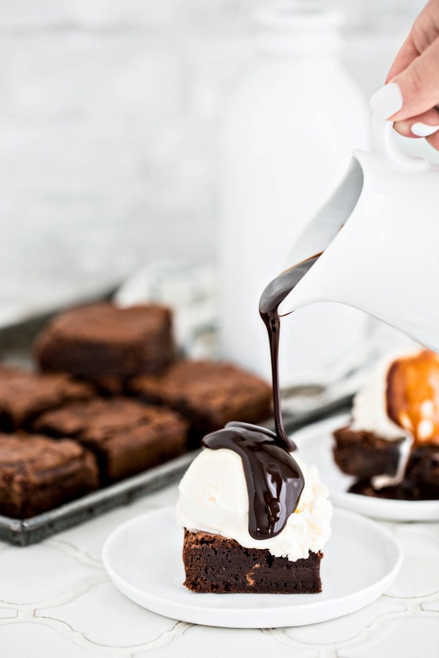 brownie on a white plate with a dollop of vanilla ice cream on top and a hand pouring a spoonful of ...