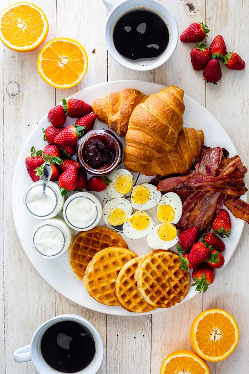 Tray of various breakfast foods including waffles, bacon, fruit, and eggs surrounded by cups of coff...