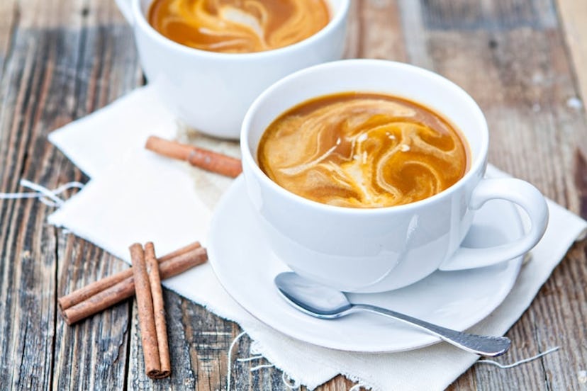 Two white mugs of caramel-colored coffee on white plates with spoons and cinnamon sticks surrounding...