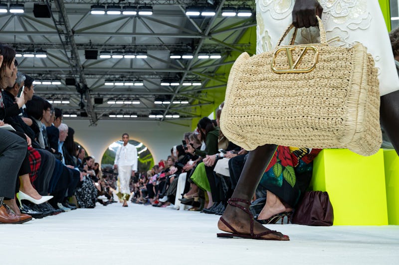 A closeup of a model walking off the runway with a woven bag