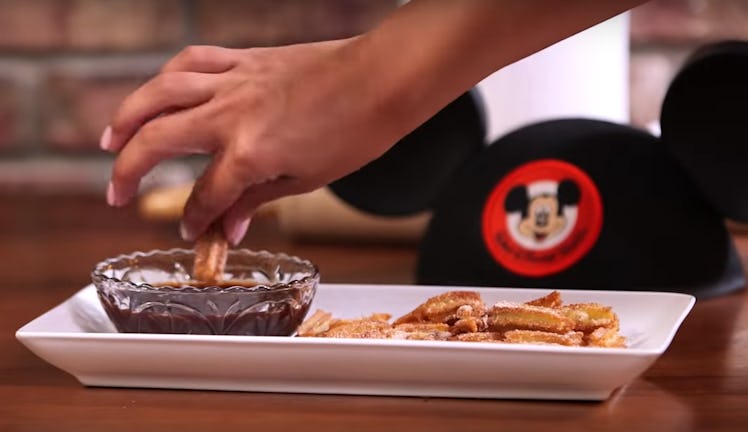 A woman's hand dips a Disney churro tot into chocolate sauce, with a Mickey Mouse hat in the backgro...