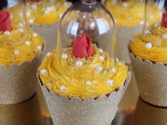 Five 'Beauty and the Beast'-themed cupcakes with red roses and yellow icing are placed on a cupcake ...