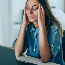 A person frowns as she rubs her temples, staring at her computer. Stress can impact your sleep, whic...