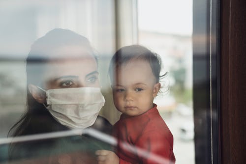 Mother in a protective facemask holds her child in front of a window