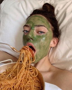 A woman with a green face mask, lying in her bed while eating spaghetti as a form of self-care