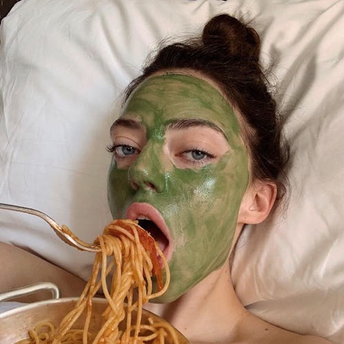 A woman with a green face mask, lying in her bed while eating spaghetti as a form of self-care