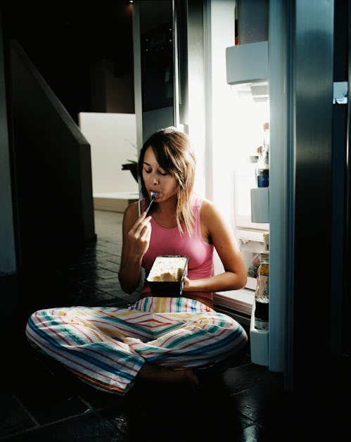 Woman in pajamas sits in front of open freezer in dark kitchen eating ice cream out of carton with a...