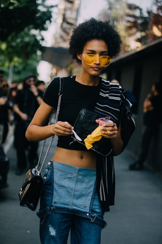 A girl wears casual half-on overalls, a black crop top, and pair of oversized yellow shades.