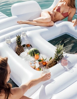 Two girls lounge on FUNBOY's Giant Dayclub pool float that has a cooler filled with drinks and fresh...