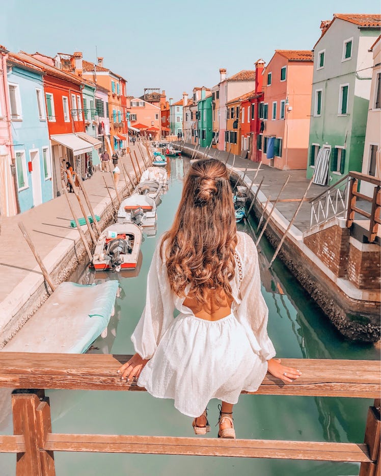 A brunette woman in a white sundress sits on a bridge overlooking a canal and colorful buildings in ...