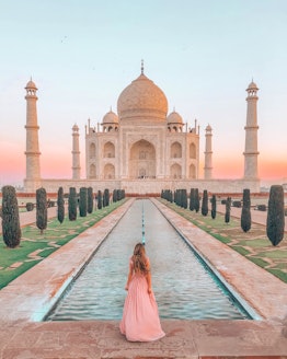 A woman in a pink maxi gown stands in front of a small pool in front of the Taj Mahal at sunset.