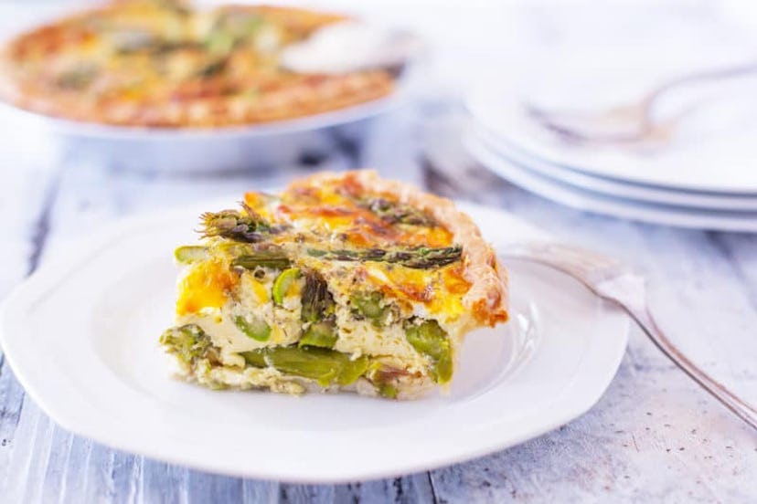 White plate with a piece of asparagus quiche and a fork on a table with casserole dish out of focus ...