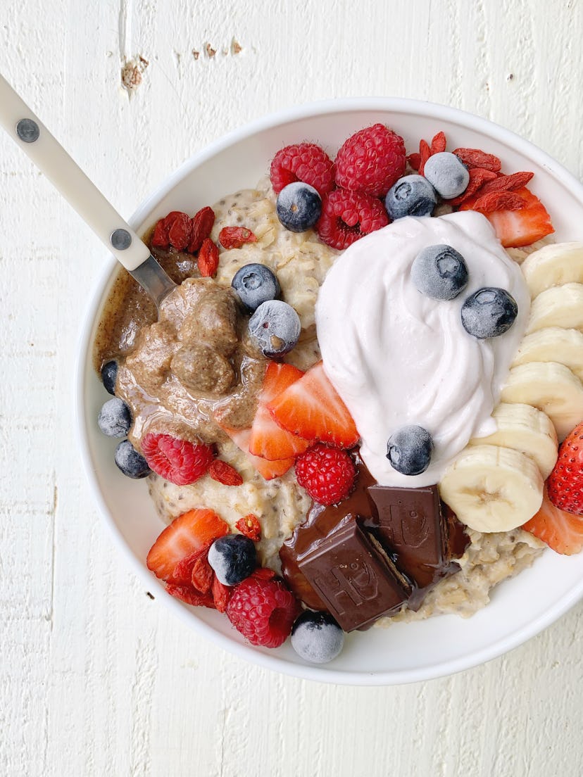 Bowl of oatmeal with berries, chocolate, yogurt, bananas, and a spoon sitting on a white table