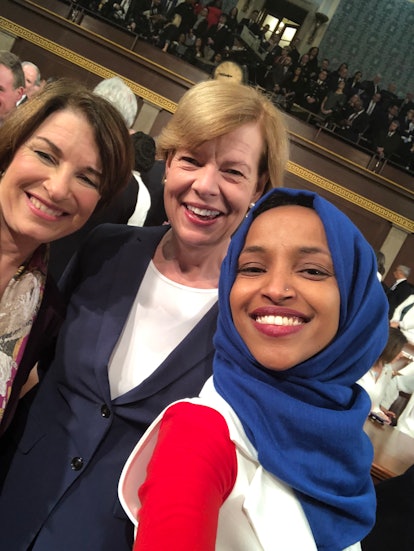 Ilhan Omar in a red and white dress and blue hijab posing with a blonde and brunette woman for a sel...