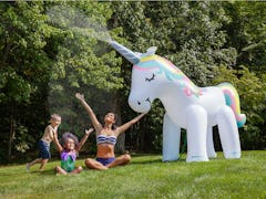 A young family sits in their backyard and plays under a giant unicorn sprinkler.