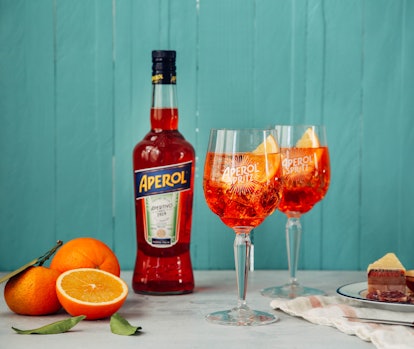 The ingredients for an Aperol Spritz sit on a table with a teal wall in the background.