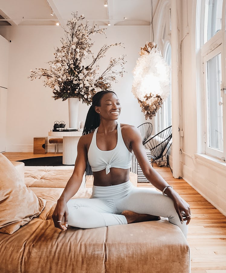 A woman in white yoga apparel smiles and sits on a brown couch in a bright room.