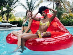 Two friends sit on a clear red lips inflatable couch while chilling in the pool on a summer day. 