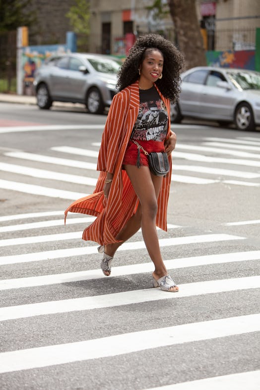 A curly girl crossing the street while wearing an orange kimono, matching shorts and a band tee.