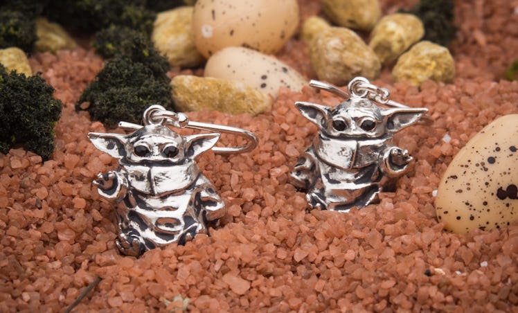A pair of sterling silver baby Yoda earrings from 'Star Wars' sits on top of brown gravel.