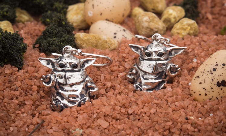 A pair of sterling silver baby Yoda earrings from 'Star Wars' sits on top of brown gravel. 