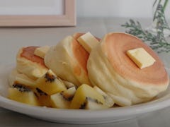 Three Japanese soufflé pancakes are on a plate with butter on top and fruit on the side. 