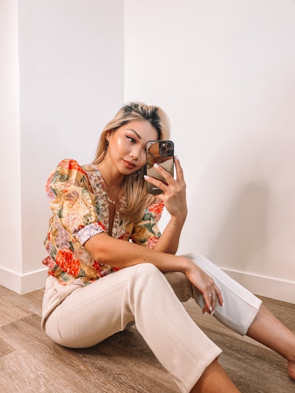 A woman with a floral top and tan capris sits on the floor and takes a mirror selfie.