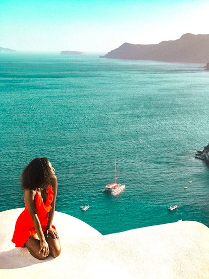 A woman in a bright red dress sits on a white stoop looking at the turquoise ocean water below.