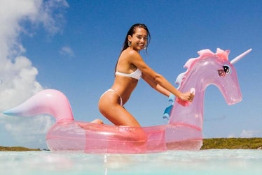 A woman sits on a pink unicorn pool float in the water. 