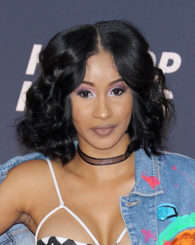 Cardi B with a curly bob parted down the middle, nude lipstick and eyeshadow in different hues of pu...