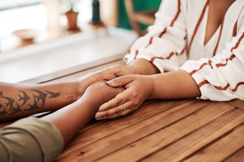 Two women hold hands on a wooden table. How To Handle Emotional Burnout As You Keep Pushing For Anti...