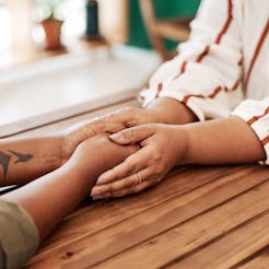 Two women hold hands on a wooden table. How To Handle Emotional Burnout As You Keep Pushing For Anti...