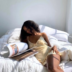 Model in a yellow slip dress flips through a book while in bed.
