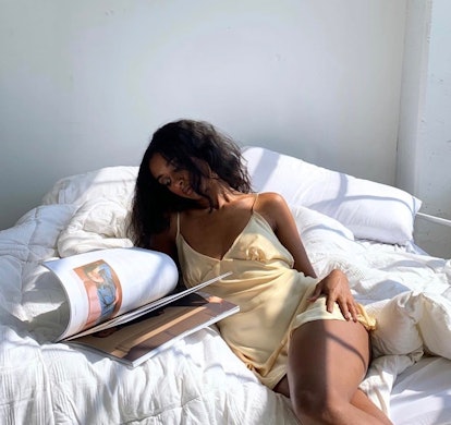 Model in a yellow slip dress flips through a book while in bed.
