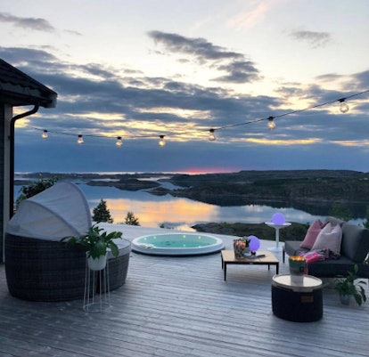 A patio overlooking a body of water has a circular hot tub and plush chairs.