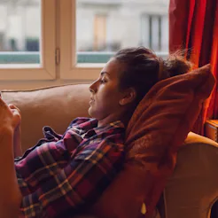 A girl in a tartan shirt lying on a couch next to a window while typing on her phone