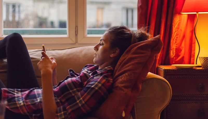 A girl in a tartan shirt lying on a couch next to a window while typing on her phone