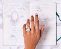 A hand with a large engagement ring on it hovers over a notebook with designs for engagement rings