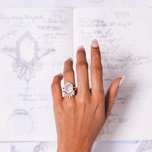 A hand with a large engagement ring on it hovers over a notebook with designs for engagement rings