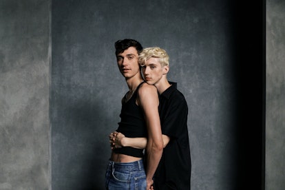 Two male models hugging in black shirts in front of a grey wall in order to promote mac cosmetics ne...
